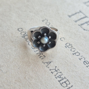 May include: A silver ring with a flower design. The ring features a flower with six petals and a small pearl in the center. The ring is set against a vintage paper background.