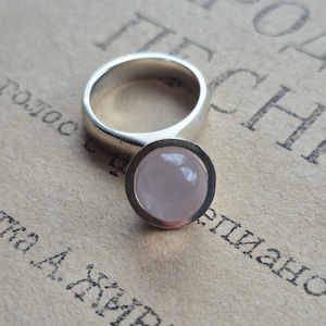 May include: A silver ring featuring a smooth, round, pale pink cabochon gemstone set in a circular bezel. The band is polished silver with a consistent width, and the stone has a soft, translucent appearance with a glossy, reflective surface.