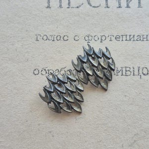 May include: Two silver-toned metal decorative elements, each composed of multiple curved, leaf-like shapes. The objects are set against a backdrop of aged paper with faded text, suggesting a vintage or antique aesthetic.
