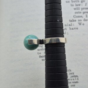 May include: A silver ring with a round, turquoise-colored stone. The ring is displayed on a black ring sizer. The background is a page from a book with visible text.