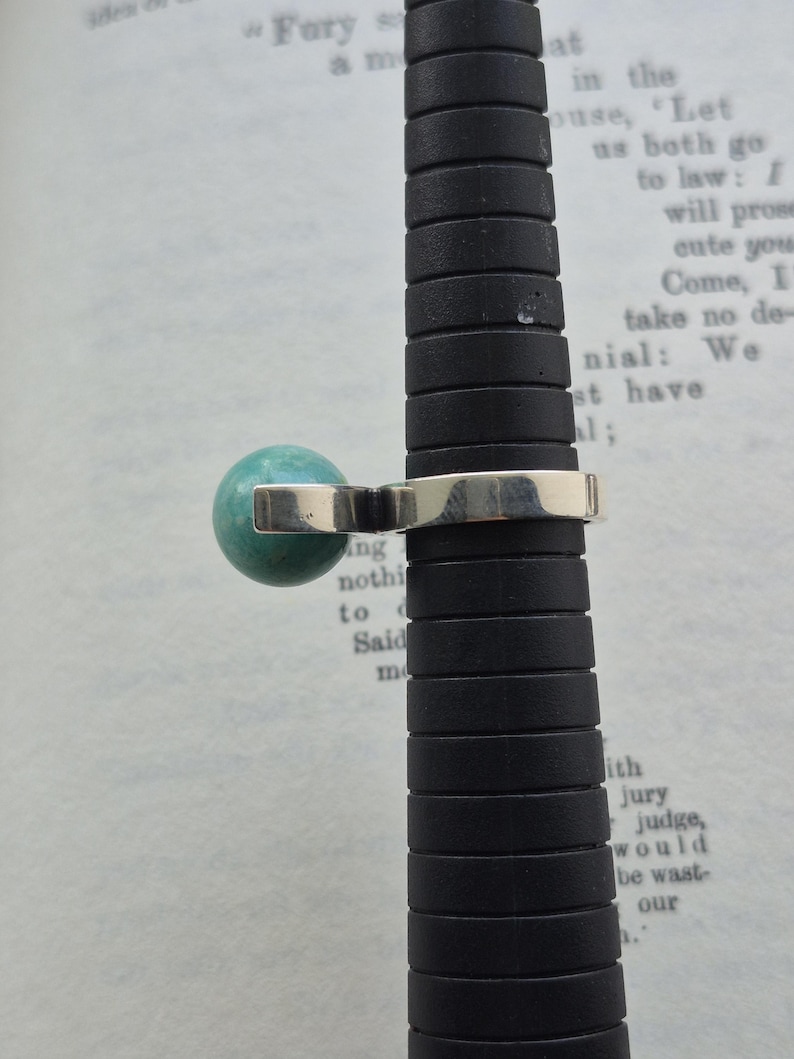 May include: A silver ring with a round, turquoise-colored stone. The ring is displayed on a black ring sizer. The background is a page from a book with visible text.