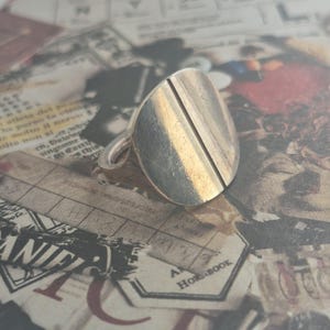 May include: A silver ring with a wide, oval-shaped band. The band has two parallel lines running down the centre.