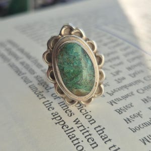 Op de afbeelding: Een zilveren ring met een ovale, groene steen in een geschulpte zetting. De ring ligt op een open boek. De steen heeft een gevlekt uiterlijk met tinten groen en bruin.