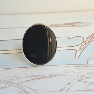 Puede incluir: Un anillo con una piedra negra grande, lisa y de forma ovalada, engastada en una banda fina de metal pulido en tono plateado. La piedra tiene una superficie brillante y reflectante que capta la luz, creando un reflejo sobre su centro oscuro.