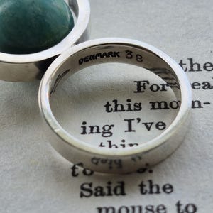 May include: A close-up of a silver ring with the inscription "DENMARK 38" inside. The ring is on a page with printed text. A silver pendant with a green stone is in the background.