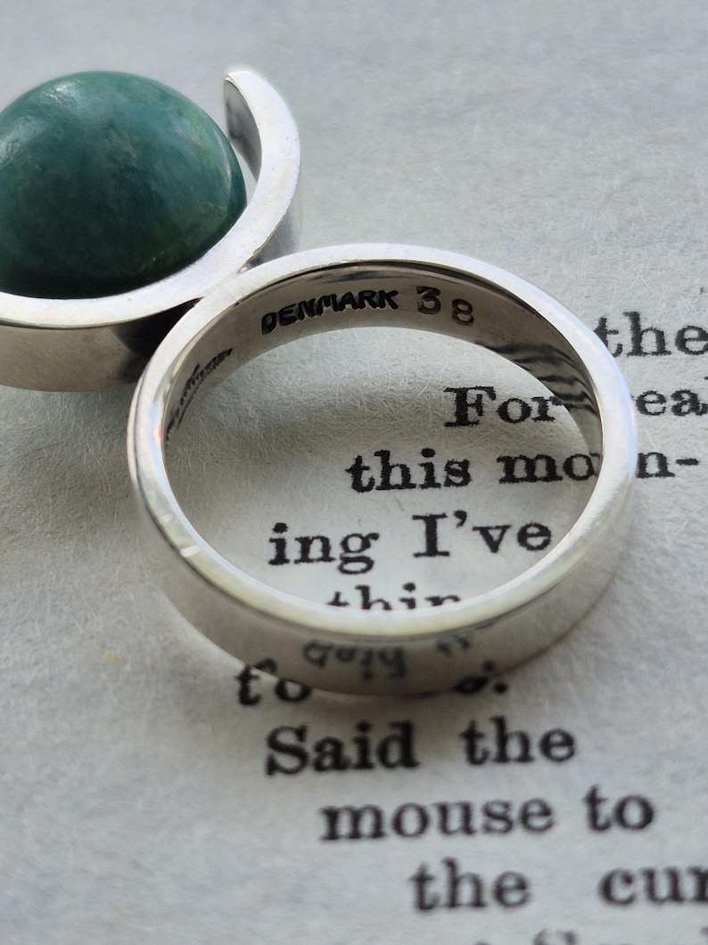 May include: A close-up of a silver ring with the inscription "DENMARK 38" inside. The ring is on a page with printed text. A silver pendant with a green stone is in the background.