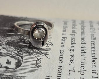 May include: A silver ring with a distinctive design. It has a clear, round stone in an oval setting. The ring is placed on a book page with text and illustrations visible. The ring's design is eye-catching.