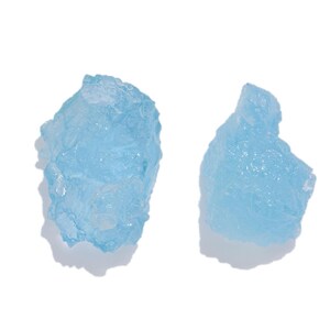 Peut inclure: Deux spécimens minéraux bruts et non polis d'une couleur bleu clair translucide. Chaque pièce présente une forme irrégulière et dentelée avec une surface cristalline texturée qui reflète la lumière, révélant des facettes naturelles.