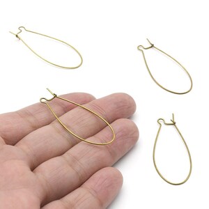 Op de afbeelding: Vier goudkleurige oorbelonderdelen in de vorm van een druppel. Eén wordt in een hand gehouden, terwijl de andere drie verspreid liggen op een wit oppervlak. De oorbellen hebben een eenvoudig, elegant ontwerp.