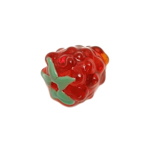 Puede incluir: Una cuenta de vidrio roja en forma de frambuesa con hojas verdes. La cuenta tiene una superficie texturizada y está sobre un fondo blanco. El objeto es decorativo.