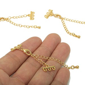 Puede incluir: Extensiones de collar doradas con cierre de mosquetón y cadena. Las extensiones tienen un diseño de tres bucles en cada extremo. Una extensión se sostiene en una mano, mientras que otras dos se muestran por separado. Hallazgos de joyería.