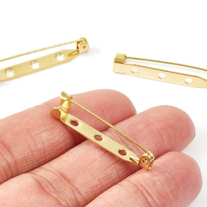 Puede incluir: Primer plano de alfileres dorados con tres agujeros para sujetar a la tela. Los alfileres tienen un cierre de seguridad. Estos alfileres se utilizan para manualidades y la fabricación de joyas.