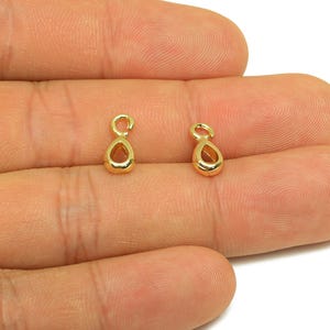 May include: Two small, gold-colored teardrop-shaped jewelry findings. Each finding has a loop at the top and a hollow, open teardrop shape. The findings are displayed against a skin-toned background.