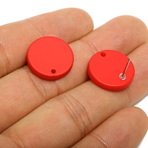 May include: Two red, round stud earrings. One earring shows the post and backing. The earrings are held in a hand, with the post of one earring visible. The earrings are a solid, bright red color.