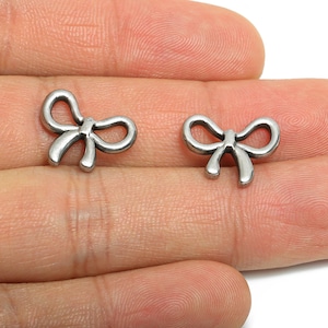 Könnte beinhalten: Zwei kleine, silberfarbene Schleifenanhänger. Die Anhänger sind aus glänzendem Metall gefertigt und haben ein schlichtes, elegantes Design. Sie werden wahrscheinlich für die Schmuckherstellung oder andere Bastelprojekte verwendet.