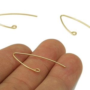 Pode incluir: Ganchos de brincos dourados. Os ganchos têm um design simples e curvo com uma extremidade em laço. A imagem mostra três ganchos, um deles segurado na mão. Adequado para fazer joias.