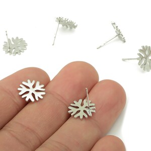 May include: Silver-colored snowflake stud earrings. The earrings have a delicate, openwork design, resembling a snowflake. Several earrings are scattered across a white background, with some held in a hand.