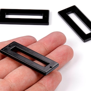 May include: Rectangular black resin jewelry findings with a hollow center. One finding is held in a hand, while two others are on a white surface. These findings have a glossy finish and small holes at each end.