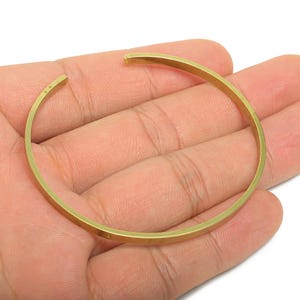 May include: A gold-coloured, open cuff bracelet. The bracelet has a simple, minimalist design with a flat, rectangular profile. The open ends allow for easy adjustment and wear. The bracelet is held in a person's hand.