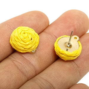 May include: A pair of yellow woven stud earrings. Each earring has a round, woven design in a bright yellow colour. The earrings have a gold-coloured post and backing. The earrings are held in a person's hand.