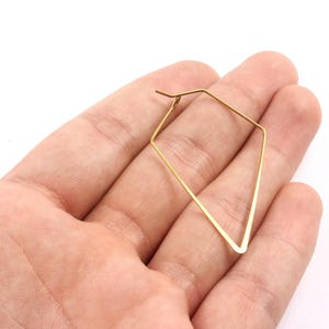 Può includere: Un singolo orecchino geometrico color oro a forma di diamante. L'orecchino è tenuto nella mano di una persona su sfondo bianco. L'orecchino è fatto di metallo sottile.