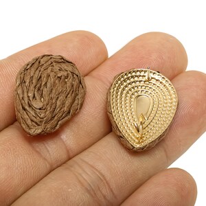 May include: Two teardrop-shaped pendants. One is a brown, textured, woven material. The other is gold-coloured with a detailed, layered design. The pendants are held in a hand against a white background.