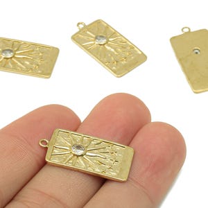 May include: Gold-colored rectangular charms with a sunburst design and a clear crystal center. The charms have a loop at the top for attaching to jewelry. The charms are made of metal and have a textured surface.