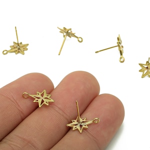 May include: Gold-coloured starburst stud earrings with a post and loop design. The earrings are small, with a detailed star shape. Several earrings are scattered on a white background, with some held in a hand.