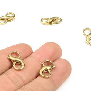 May include: Several gold-colored lobster claw clasps. The clasps are in the shape of an infinity symbol. The clasps are made of metal and are on a white background. Some clasps are held in a hand.
