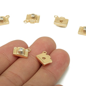 May include: Gold-colored camera charms with a small, clear gem on the lens. The charms are small, with a loop at the top for attaching to jewelry. The camera charms are shown in a hand against a white background.