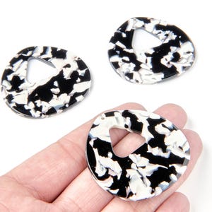 Peut inclure: Trois pendentifs en résine marbrée noire et blanche avec un design triangulaire ouvert unique. Les pendentifs ovales ont un trou central et sont tenus dans une main sur fond blanc. La résine présente un motif moucheté.