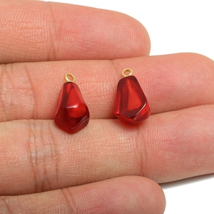 Könnte beinhalten: Zwei rote, tropfenförmige Anhänger mit goldfarbenen Schlaufen. Die Anhänger haben eine durchscheinende, tiefrote Farbe. Die Anhänger werden in einer Hand vor weißem Hintergrund gehalten.