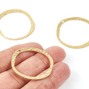 Puede incluir: Tres colgantes ovalados de metal dorado con textura martillada y pequeños agujeros para hacer joyas. Los colgantes tienen una forma irregular y se muestran sobre un fondo blanco.
