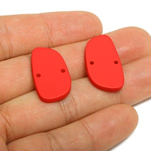 Pode incluir: Dois componentes vermelhos brilhantes, em forma oval, com bordas arredondadas e dois pequenos orifícios. Estes componentes são provavelmente para fazer joias ou artesanato. Os componentes são mostrados em um fundo neutro.