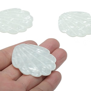 May include: Three translucent, white seashell-shaped craft embellishments. One is held in a hand, showcasing the detailed, ridged texture. The other two are positioned above. These decorative pieces are ideal for jewelry making or other craft projects.