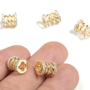 May include: Gold-colored metal jewelry beads with a spiral design and clear crystal accents. The beads are cylindrical with a series of loops and are shown on a white background. The beads are small, likely for jewelry making.
