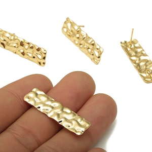 May include: Gold-coloured rectangular earrings with a hammered texture. The earrings are shown in a studio setting against a white background. The earrings are approximately 2.5 cm long.