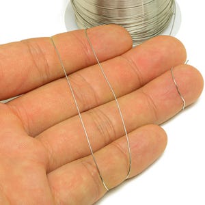 May include: Silver-colored craft wire is displayed, with strands held against a hand. A spool of the wire is visible in the background. The wire appears thin and flexible, suitable for jewelry making or other crafts.