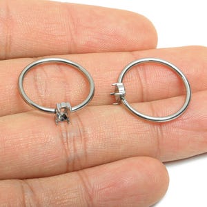 Könnte beinhalten: Zwei silberfarbene Ringrohlinge mit Krappenfassung für einen Edelstein. Die Ringe sind rund und werden in der Hand einer Person gehalten. Die Krappenfassungen sind für einen quadratischen oder rechteckigen Stein konzipiert.