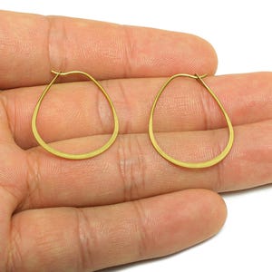 May include: A pair of gold-colored teardrop-shaped hoop earrings. The earrings are displayed on a hand, showcasing their delicate design and warm metallic hue. The hoops have a simple, elegant style.