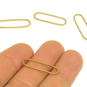 May include: Close-up of several small, gold-colored oval-shaped metal jewelry findings. One is held in a hand, with others scattered around. The findings have a simple, open design, suitable for jewelry making or crafting projects.