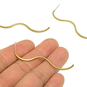 May include: Gold-colored, wavy earrings with a post backing. The earrings have a flowing, organic shape, resembling a stylized wave. The earrings are shown against a white background.