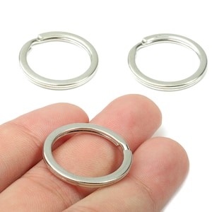 Puede incluir: Llaveros divididos de color plateado. Un anillo se sostiene en una mano, con dos anillos adicionales visibles en el fondo. Los anillos son de metal y tienen un diseño dividido para sujetar llaves u otros objetos.