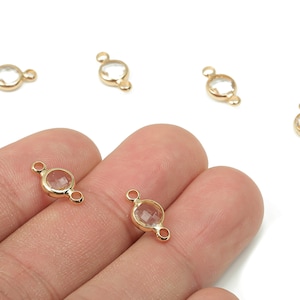 May include: Gold-colored jewelry findings with clear, round, faceted stones. The findings have a circular stone setting with a loop on each side for attaching to other jewelry components. Several pieces are scattered on a white background.