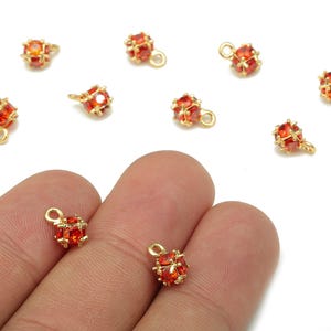 May include: Gold-colored metal charms with a small, round, cage-like design, each encasing a cluster of orange-red gemstones. The charms have a small loop for attaching to jewelry. Several charms are scattered on a white background.