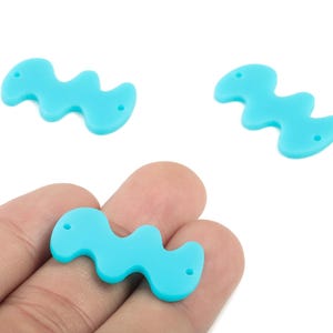 Könnte beinhalten: Drei hellblaue, wellenförmige Anhänger mit jeweils zwei Löchern. Einer der Anhänger wird in einer Hand gehalten. Die Anhänger bestehen aus einem glatten Material, für Schmuckherstellung oder Bastelarbeiten.
