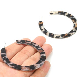 May include: Two hoop earrings with a leopard print pattern. The earrings are a circular shape with a post for piercing. The print features black, brown, and tan spots on a clear background. One earring is held in a hand.