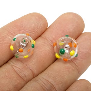 May include: Two clear glass beads with colourful polka dots in orange, green, and yellow. The beads are round and have a hole through the centre. The beads are held in a person's hand.