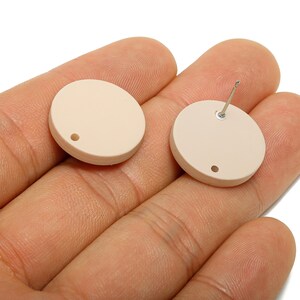 May include: Two beige, circular earrings. One earring has a post attached, while the other has a small hole. The earrings are shown against a neutral background.