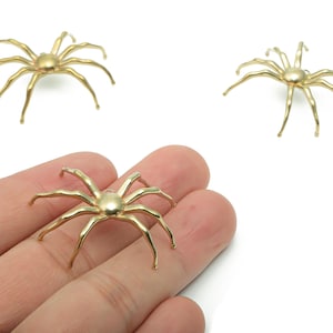 May include: Gold-colored spider charms with eight legs and a central body. The charms are shown in a group of three, with one held in a hand. These charms are suitable for jewelry making or craft projects.
